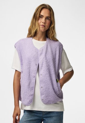 Femme aux longs cheveux blonds porte un gilet texturé lavande boutonné par-dessus un T-shirt blanc et un jean bleu, debout avec une main dans la poche.