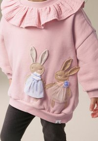 Sweatshirt rose clair avec un col froncé, présentant deux lapins brodés en robes. Les lapins ont des détails beiges et pastel.