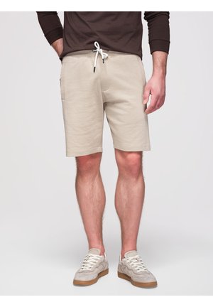Mann trägt beige Shorts mit Kordelzug, hellbeige Sneakers und ein dunkles Langarmshirt und steht vor einem einfarbigen Hintergrund.
