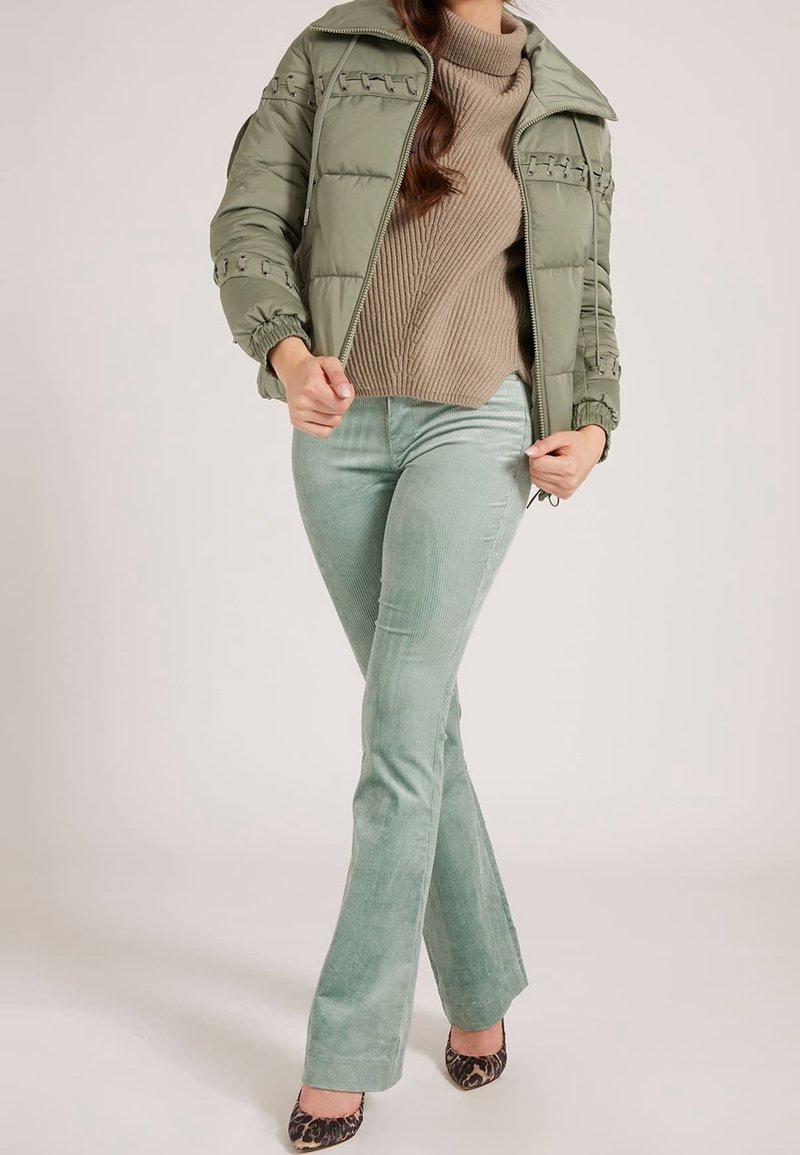 Femme portant un pantalon évasé en velours côtelé vert clair, une doudoune vert olive, un pull à col roulé côtelé beige et des talons imprimés léopard.