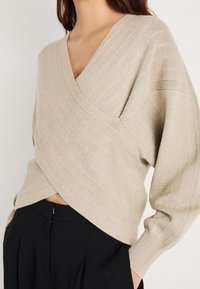 Femme portant un pull beige en maille côtelée style cache-cœur à manches longues et un pantalon noir taille haute devant un fond uni.