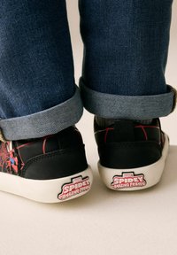 Mustat lenkkarit, joissa on punainen hämähäkinseittimäinen kuvio, siinä on Marvelin Spidey -graafikko kantapäällä, valkoinen pohja ja farkkupaikat.