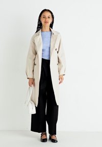 Beige trench kabát hosszú ujjakkal, dupla gombolású elülső résszel, csíkos kék és fehér ing fölött, fekete, bő szárú nadrággal és lapos cipővel párosítva.