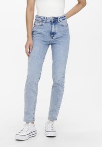 Personne portant un jean skinny taille haute bleu clair et des baskets blanches, debout devant un fond uni.