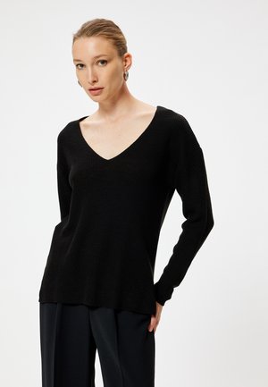 Schwarzer Strickpullover mit tiefem V-Ausschnitt, langen Ärmeln und lockerer Passform, kombiniert mit lockeren schwarzen Hosen, vor einem schlichten Hintergrund.