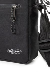 Zaino Eastpak nero con forma rettangolare, tessuto testurizzato, tasca con zip, dettagli in gomma e tracolla regolabile. Logo visibile sulla parte anteriore.
