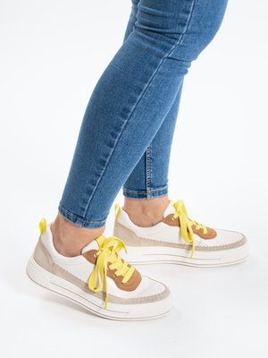 Legs in blue skinny jeans wearing white sneakers with beige and brown accents and yellow laces, standing on a white background.