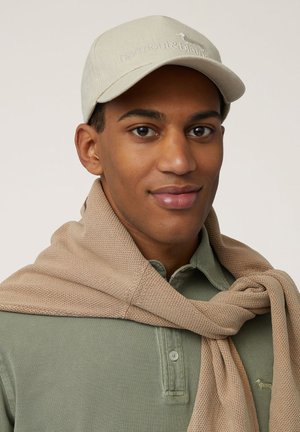 Beige cap with logo, green polo shirt with buttons, and beige textured scarf draped casually around the neck. Skin tone appears neutral.