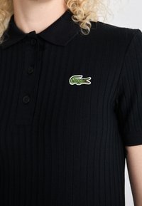 Personne portant un polo côtelé noir avec un logo crocodile vert brodé sur la poitrine gauche.