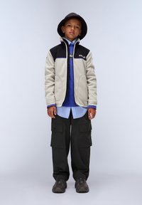 Fleecejacka med en svart övre del och beige nedre del. Blå lager-på-lager skjorta, mörka cargobyxor och svart bucket hat. Grå sneakers.