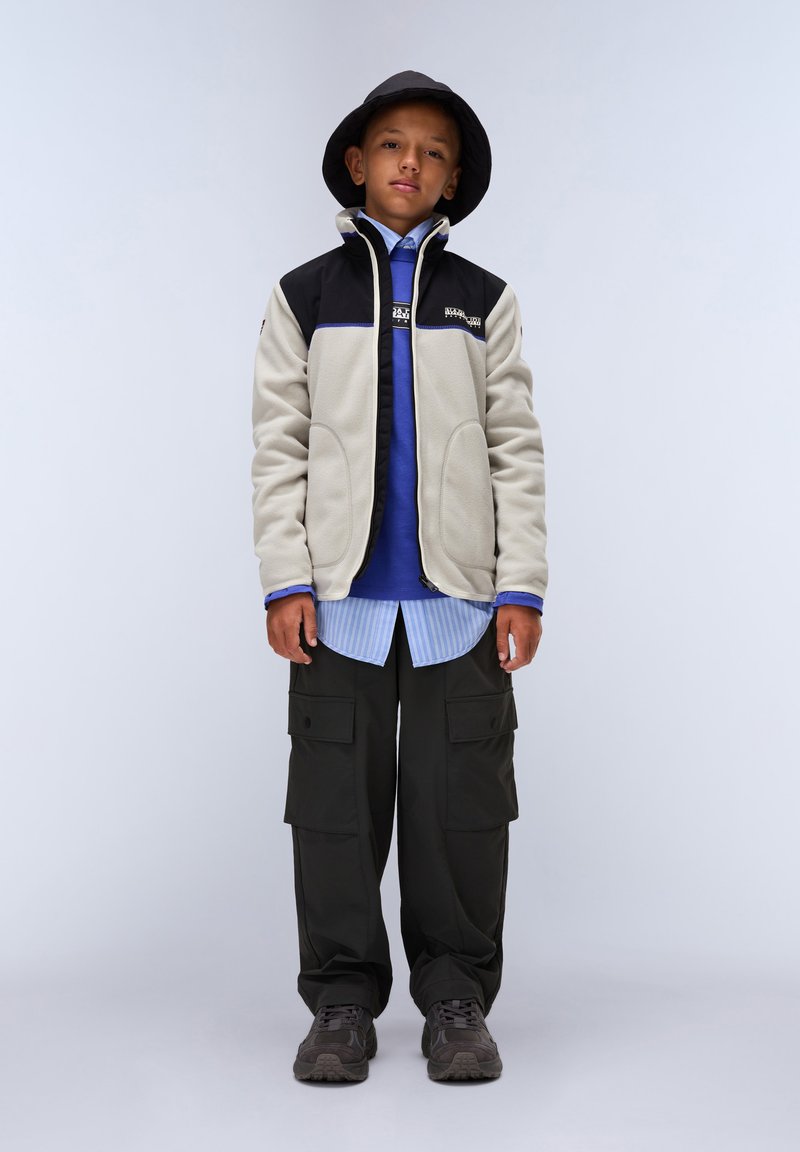 Fleecejacka med en svart övre del och beige nedre del. Blå lager-på-lager skjorta, mörka cargobyxor och svart bucket hat. Grå sneakers.