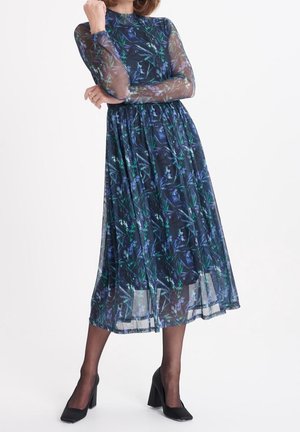 Frau, die ein langärmeliges, marineblaues Kleid mit hohem Kragen und Blumenmuster trägt, durchsichtige Ärmel, schwarze transparente Strumpfhosen und schwarze Schuhe mit Blockabsatz.