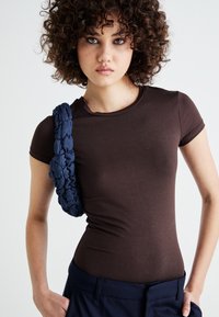 Brown fitted t-shirt made of stretchy cotton, featuring a braided navy strap across one shoulder, paired with navy trousers.