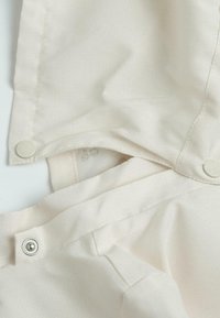 Tissu beige avec une texture lisse, doté d'une fermeture à bouton-pression. Le design présente des motifs en grille subtils et une apparence superposée.