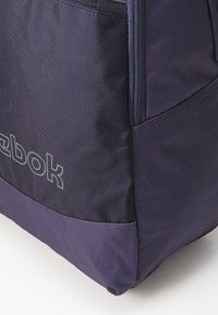 Purpurinė nailoninė maišeliai su tekstūruotu paviršiumi, turi užtrauktuko uždarymą ir ryškiai pilką Reebok logotipą, atspausdintą priekyje.
