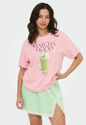 Vrouw draagt een roze "Matcha Mood" t-shirt met groene drankgrafiek en muntgroene rok met kanten rand, poseert met hand op de heup.
