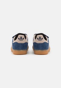 Siniset mokkakengät vaaleanruskealla kumipohjalla ja mustalla sisävuorella, joissa on vaaleanpunainen kantaosio logolla. Kaksi säädettävää remmiä näkyvissä.