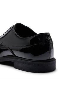 Chaussure noire en cuir verni avec un bout arrondi, finition lisse et un laçage. Présente une semelle noire texturée et un détail de logo en relief sur le talon.