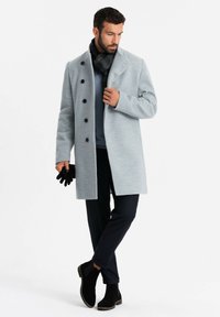 Abrigo gris claro con un alto cuello, botones negros, combinado con un suéter azul, guantes negros y pantalones oscuros, complementado con botas negras hasta el tobillo.