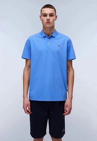 Young man standing, wearing a light blue short-sleeve polo shirt and black shorts, against a plain light background.