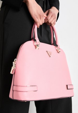 Mains tenant un sac à main structuré rose avec des ferrures dorées sur une tenue noire et un fond uni.