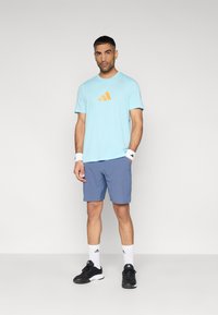 Ανοιχτό μπλε αθλητικό μπλουζάκι με πορτοκαλί λογότυπο, σετ με μπλε σορτς· λευκά βραχιόλια καρπού και κάλτσες· μαύρα αθλητικά παπούτσια. Απλός, λειτουργικός σχεδιασμός.