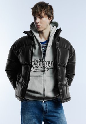 Črna puhasta jakna z bleščečo teksturo, oblečena čez sivo kapuco z vezenim napisom, kombinirana z modrimi kavbojkami iz jeansa.