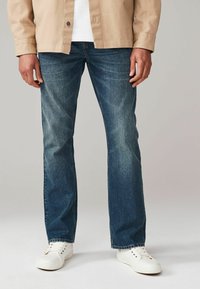 Jeans din denim de un albastru închis, cu un finisaj ușor decolorat, design cu picioare drepte, asociați cu adidași albi și o cămașă bej.