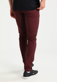 Burgunderfärgade, slim-fit byxor i bomull, med en slät textur och bakfickor. Bäraren har svarta sneakers.