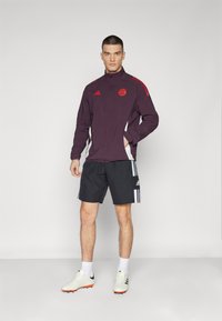 Burgunder athletic jakke med røde detaljer og hvite sidestykker, kombinert med svarte shorts med hvite striper og fotballsko.