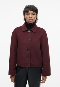 Bordeaux uldblandede jakke med krave, sorte knapper, cropped design og vide ærmer, parret med en sort turtleneck.