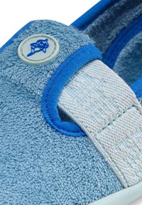 Gros plan sur une chaussure en tissu bleu texturé avec une bande élastique blanche et un bord bleu, ornée d'un petit bouton rond avec un logo bleu.