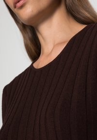 Top in maglia a coste marrone con scollatura tonda, caratterizzato da una texture verticale e una leggera lucentezza, accorciato sulle spalle.