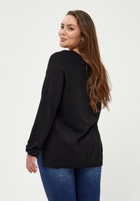 Schwarzer Pullover mit langen Ärmeln, lockerer Passform und ribbeltem Saum. Zusammen mit blauen Jeans trägt das Modell lange Haare und ist leicht zur Seite gedreht.