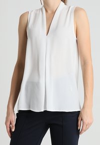 Blusa bianca senza maniche con scollatura a V, realizzata in tessuto leggero, con una vestibilità rilassata e leggermente trasparente.
