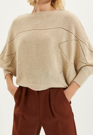 Vrouw draagt een beige losse gebreide trui met open steekdetails en roestkleurige broek met hoge taille, handen in de zakken.