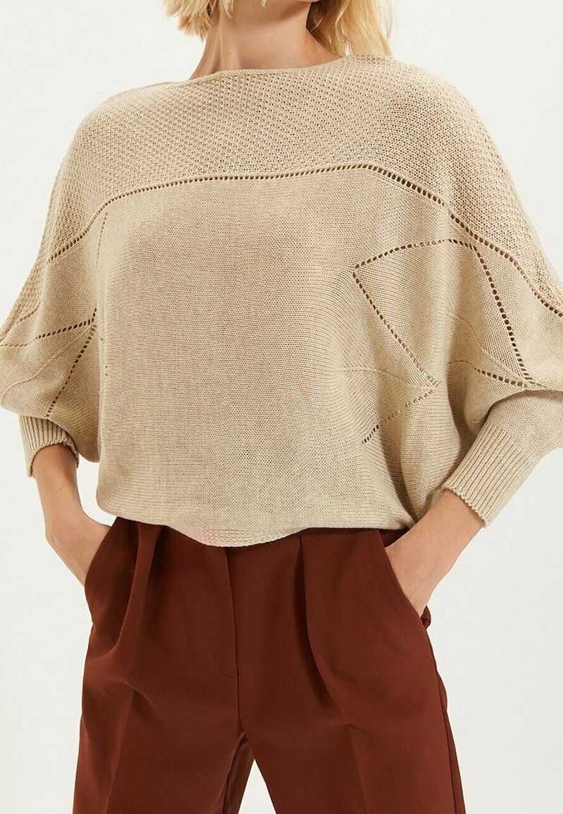 Femme portant un pull en maille beige ample avec des détails en points ouverts et un pantalon taille haute couleur rouille, les mains dans les poches.