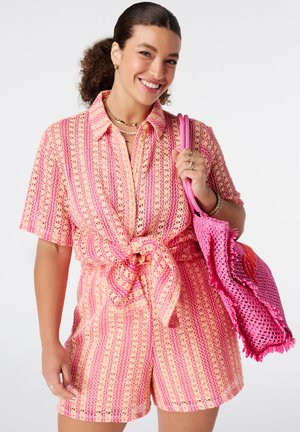 Vrouw glimlacht, draagt een roze en crèmekleurig gestreept gehaakt shirt geknoopt in de taille en bijpassende korte broek, houdt een roze geweven handtas vast.