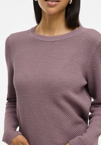 Pull en tricot de couleur mauve doux, doté d'un motif texturé, d'un col rond et de manches longues. Des poignets côtelés ajoutent une touche de détail au design.