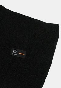 Tessuto a maglia nero con una piccola etichetta rettangolare nera che presenta un cerchio bianco, testo bianco e una linea orizzontale arancione.
