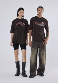 To modeller bærer oversized brune grafiske t-shirts med teksten "OFFLINE FOR PRIVATE REASONS". Den kvindelige model sætter det sammen med sorte shorts og knæ-høje støvler, mens den mandlige model har baggy forvaskede jeans og elegante sorte sko på.