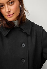 Cappotto di lana nera con ampio colletto, caratterizzato da tre grandi bottoni neri e una finitura testurizzata. Indossato sopra un dolcevita grigio.