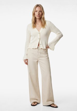 Vrouw draagt een crèmekleurig geribbelde cardigan met getande randen, beige wijde broek en bruine sandalen, staand tegen een witte achtergrond.