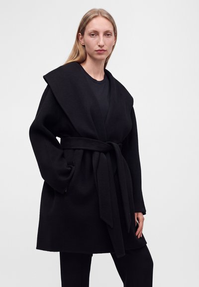 Femme blonde porte un manteau noir ceinturé avec un large col sur des vêtements noirs, debout devant un fond gris clair uni.