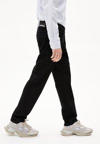 Pantalons en denim noir avec une coupe droite, associés à des baskets gris clair avec des accents prononcés. Le mannequin porte une chemise rayée bleue et blanche.