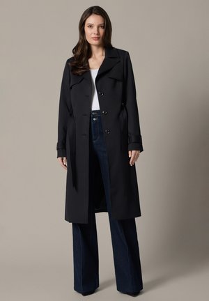 Kobieta stojąca w długim czarnym płaszczu typu trencz na białym topie i ciemnoniebieskich szerokich dżinsach na jednolitym beżowym tle.