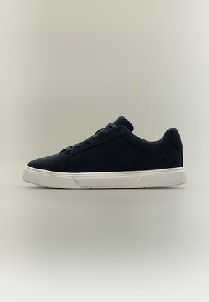 Zwarte lage sneaker met witte rubberen zool en veters, zijaanzicht op een effen lichte achtergrond.