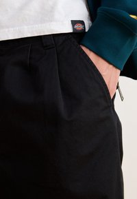 Svarta bomullsbyxor med avslappnad passform, med sidofickor och en synlig varumärkesetikett på midjebandet. Texturen är len med minimala veck.