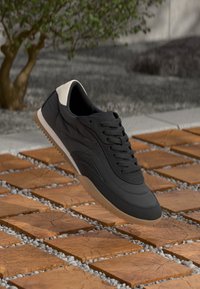 Pantof sport negru cu călcâi bej și talpă de gumă plutind deasupra unei dale de lemn și a solului cu pietre în aer liber.