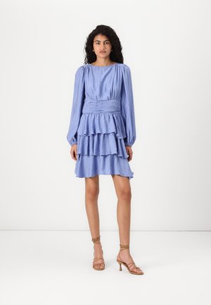 Blauwe gerimpelde jurk met lange mouwen, gathered taille en gelaagde rok. De stof heeft een subtiele glans en een gestructureerde afwerking. Gecombineerd met sandalen met bandjes.